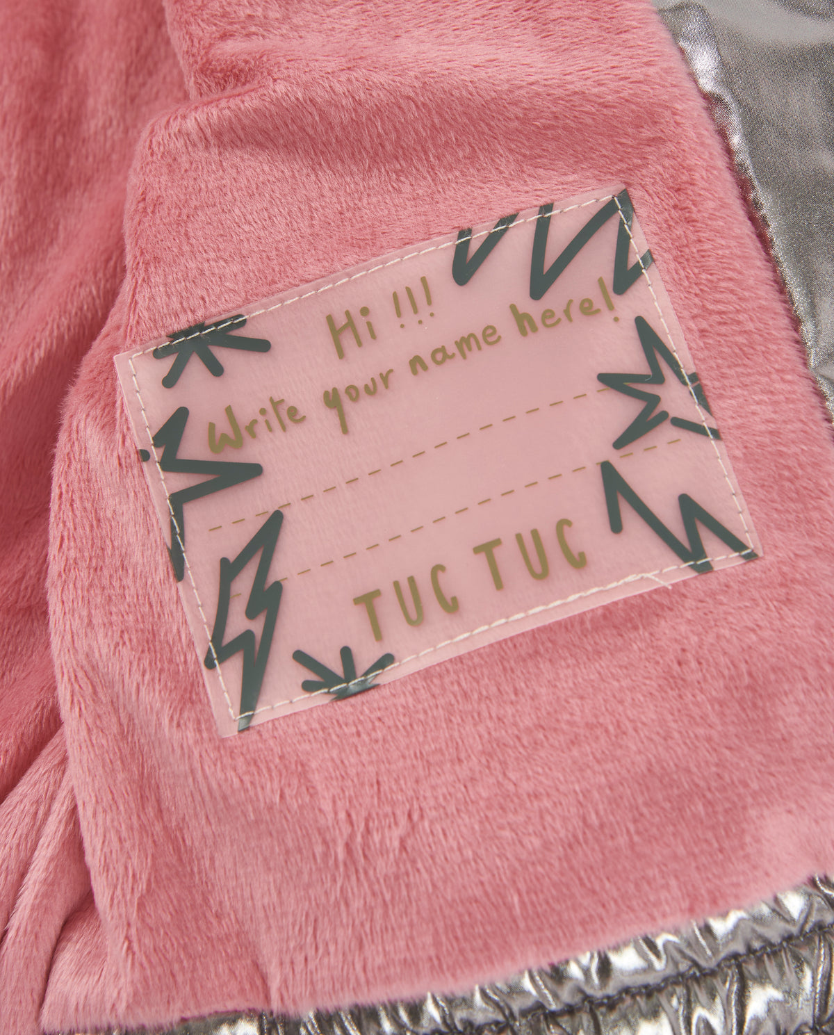 Montun pembe pelüş astarı ve 'Write your name here' yazılı Tuc Tuc isim etiketi yakın planda.