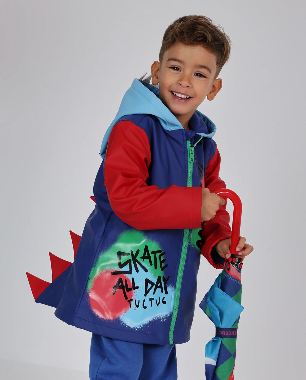 Tuc Tuc lacivert kırmızı dinozor dikenli yağmurluğu giymiş erkek çocuk, elinde şemsiyeyle; sırttaki kırmızı dikenler ve Skate All Day baskısı görünüyor.