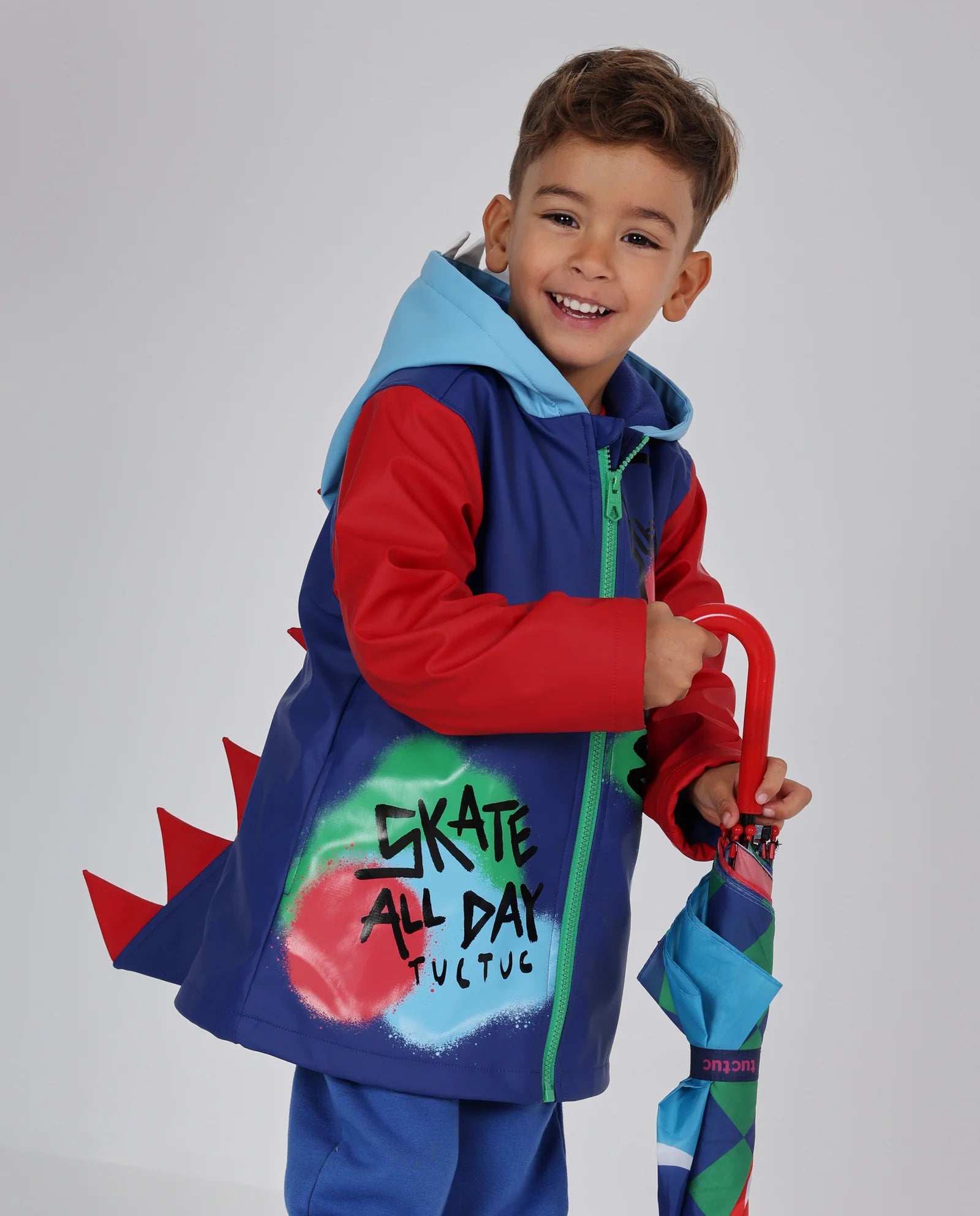 Tuc Tuc lacivert kırmızı dinozor dikenli yağmurluğu giymiş erkek çocuk, elinde şemsiyeyle; sırttaki kırmızı dikenler ve Skate All Day baskısı görünüyor.
