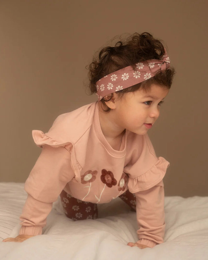 Dirkje pudra pembe sweatshirt kız çocuk üzerinde yakın planda; omuz fırfırı, çiçek nakışı ve papatyalı saç bandı.