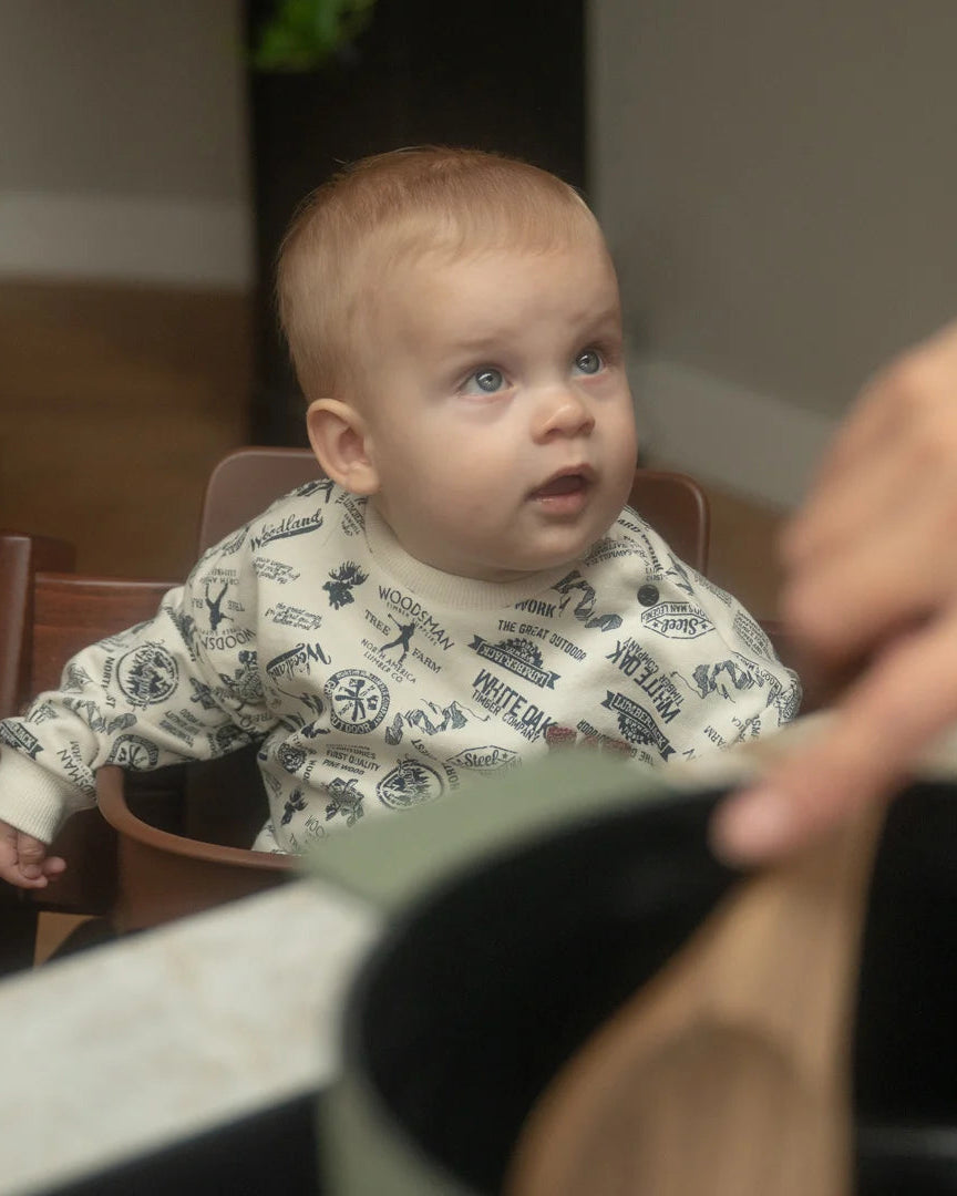 Dirkje krem woodland desenli sweatshirt bebeğin üzerinde; mutfakta mama sandalyesinde, lacivert pantolonla kombinlenmiş.