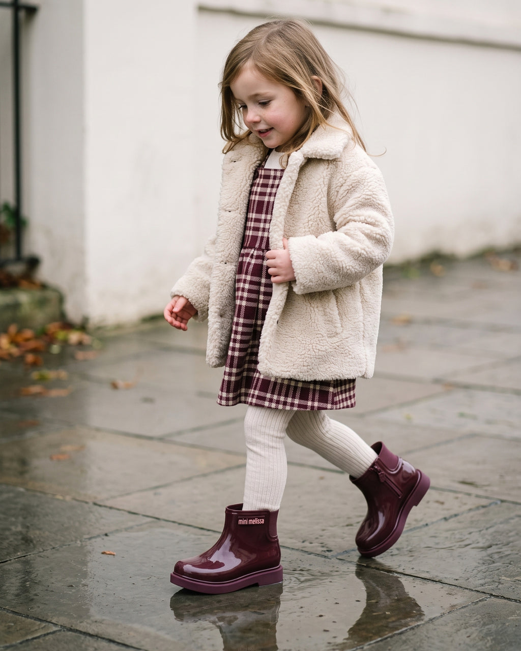 Krem peluş mont ve bordo-krem ekose elbise kombiniyle bordo Mini Melissa bot giyen kız çocuğu, yağmur sonrası ıslak kaldırımda yürürken.