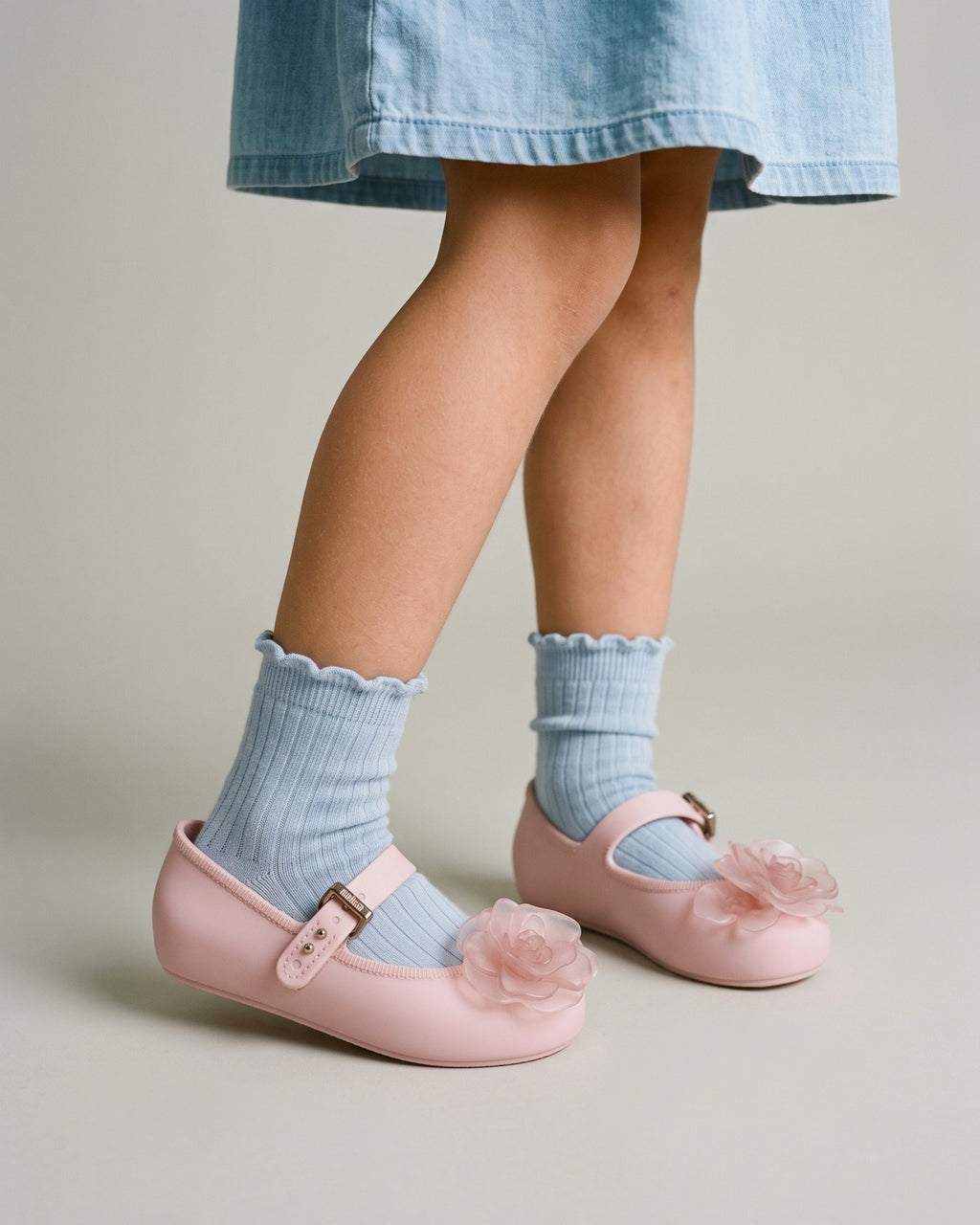 Ayakta duran kız çocuğunun ayağında pembe Mini Melissa babet ayakkabı; mavi fırfırlı soket, ince düz taban ve tokalı bant yakın planda.