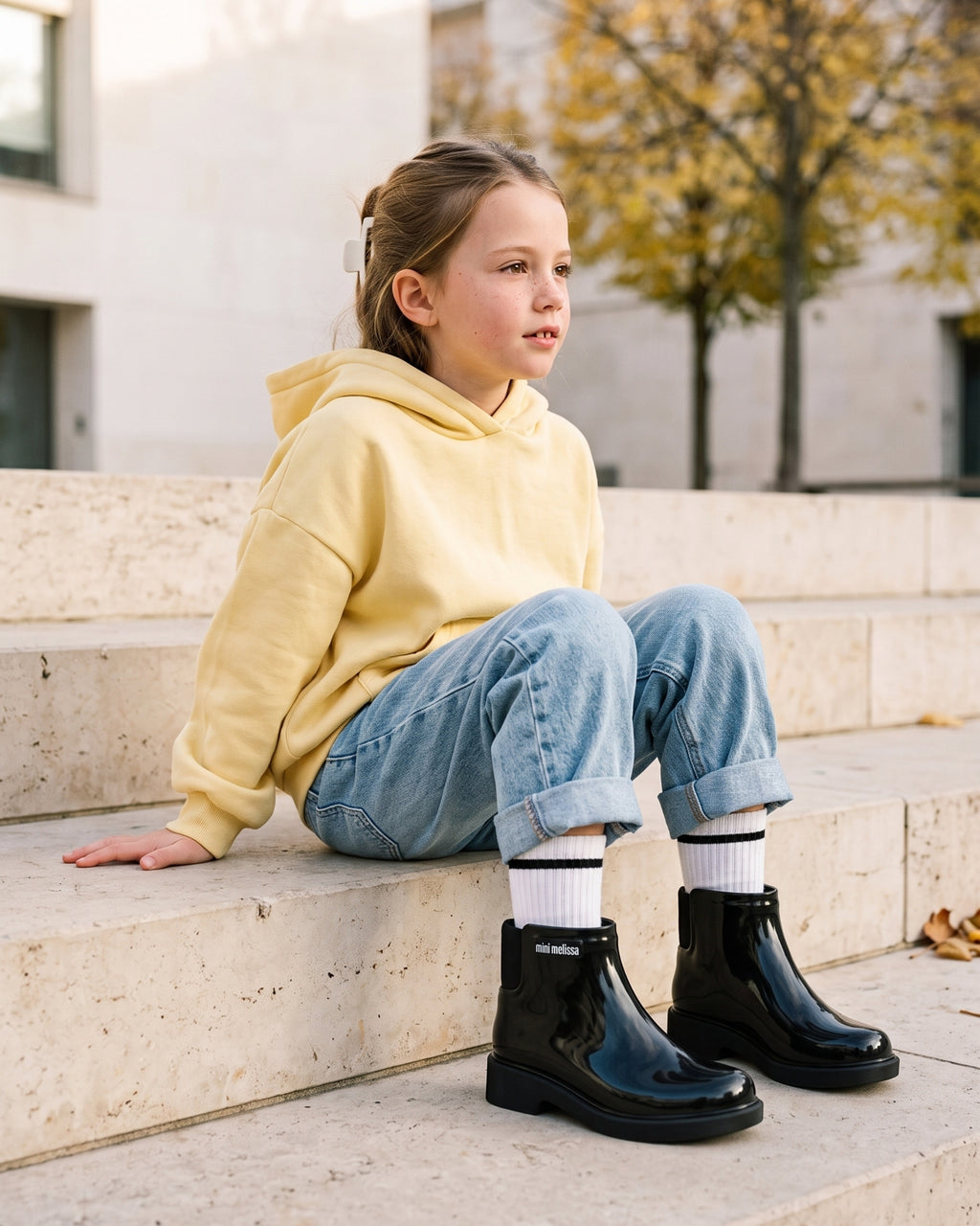 Tereyağı sarısı sweatshirt ve bol kesim kot kombiniyle siyah Mini Melissa Chelsea bot giyen okul çağındaki kız çocuğu, şehir meydanının taş basamaklarında; sonbahar ve kış için kız çocuk kışlık ayakkabısı.