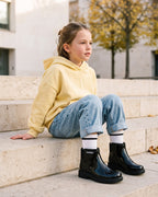 Tereyağı sarısı sweatshirt ve bol kesim kot kombiniyle siyah Mini Melissa Chelsea bot giyen okul çağındaki kız çocuğu, şehir meydanının taş basamaklarında; sonbahar ve kış için kız çocuk kışlık ayakkabısı.