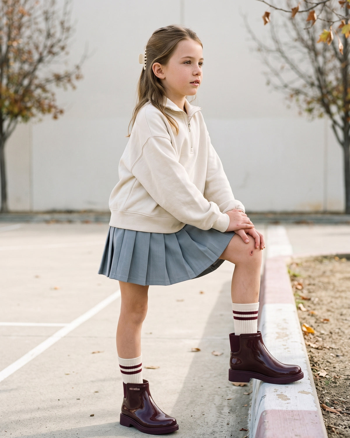 Ekru sweatshirt, pileli tenis eteği ve bordo çizgili çorap kombiniyle bordo Mini Melissa Chelsea bot giyen okul çağındaki kız çocuğu, okul bahçesinin kenarında.