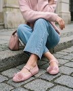 Açık mavi denim ve pembe triko kombiniyle pembe babet giyen kadın, taş kaldırımda otururken; ayaktaki gerçek oranı ve şeffaf gül aplikeyi gösteren sokak karesi.