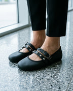 Terrazzo zemin üzerinde duran siyah Melissa Soft Ballerina Studs kadın babetlerin yakın plan görünümü; çift bant üzerindeki gümüş metal zımba detayları ve mat Melflex doku