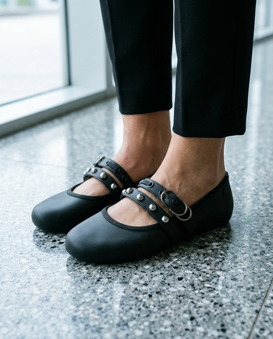 Terrazzo zemin üzerinde duran siyah Melissa Soft Ballerina Studs kadın babetlerin yakın plan görünümü; çift bant üzerindeki gümüş metal zımba detayları ve mat Melflex doku