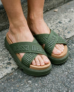 Beton zemin üzerinde duran haki yeşili Melissa Cross M-Lover Platform kadın terliklerin yakın plan görünümü; çapraz bantlar üzerinde kabartmalı Melissa logo detayı ve taba rengi konforlu taban.