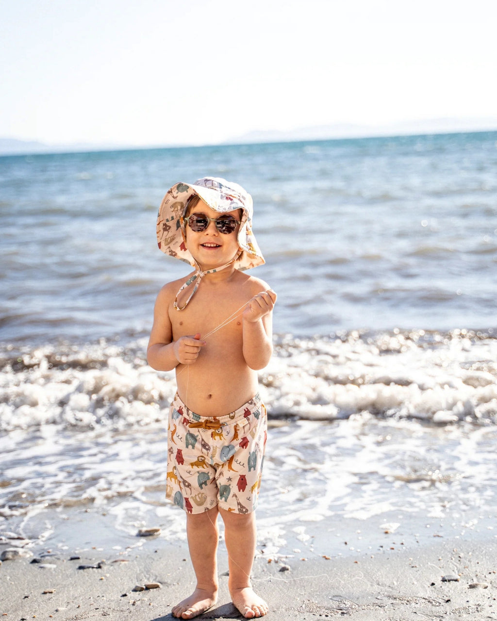 Miela Kids jungle mayo şortlu küçük erkek çocuk sahilde duruyor, şapka ve güneş gözlüğü, deniz arka plan