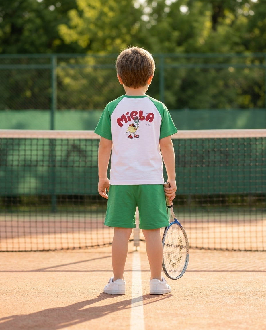 Miela Kids tennis takımlı erkek çocuk tenis kortunda arkadan, raket elinde duruyor
