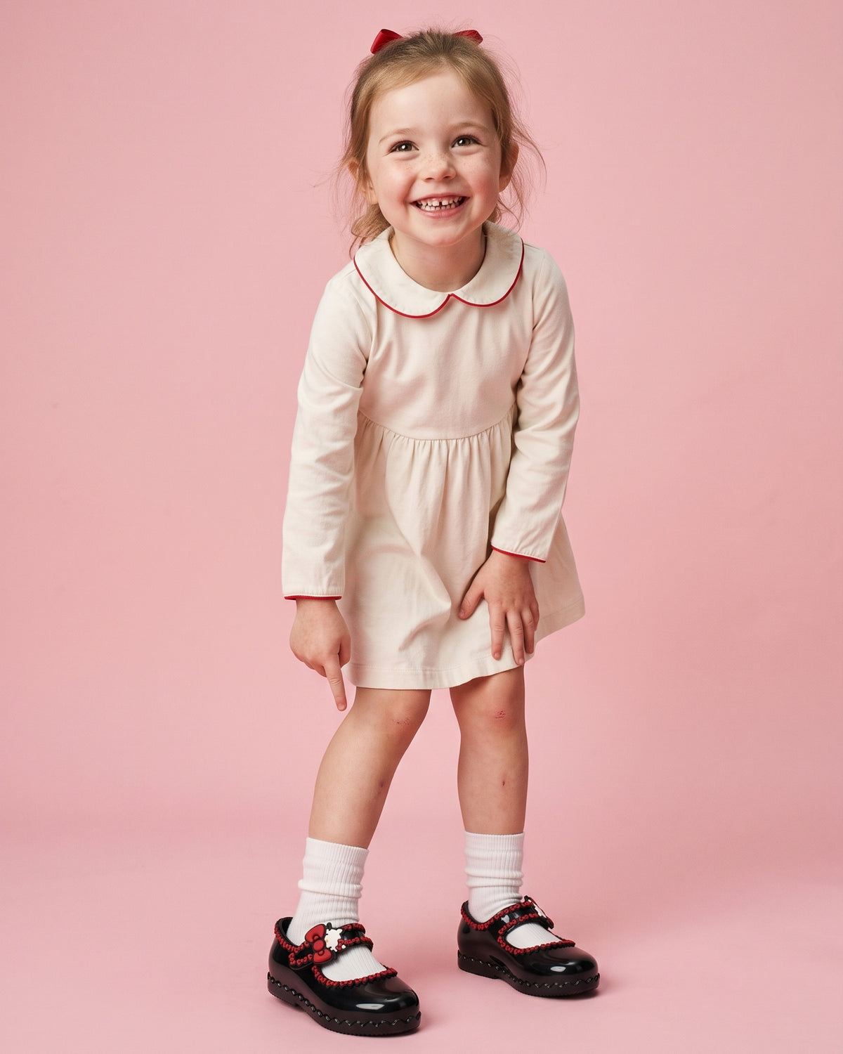 Mini Melissa Hello Kitty ayakkabı giyen kız çocuk; siyah parlak babet, kırmızı fiyonk detayları ve ekru elbise kombini.