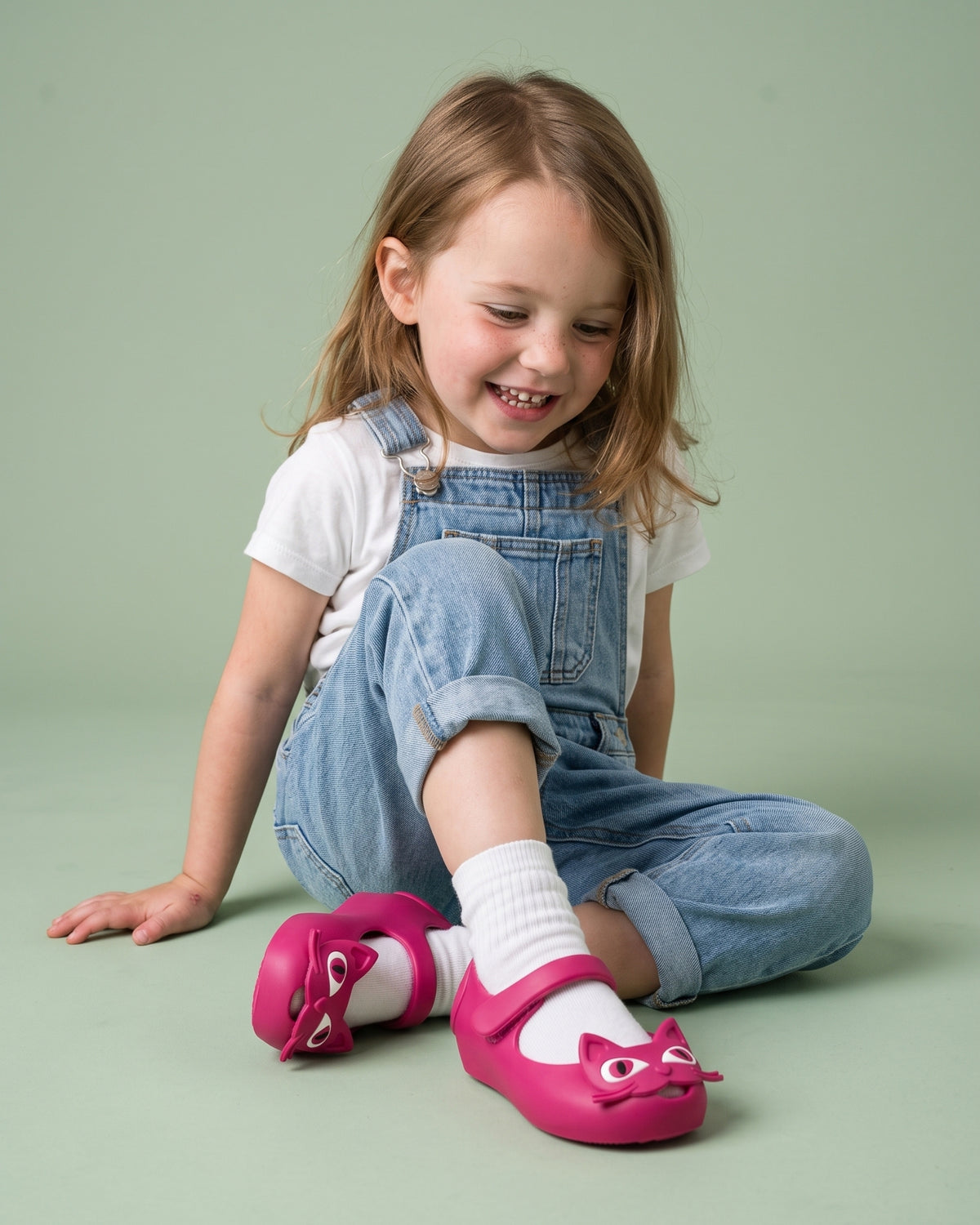 Denim salopet ve beyaz tişört kombiniyle fuşya Mini Melissa babet giyen küçük kız çocuğu, stüdyoda yerde otururken; 22-27 numara kız bebek ayakkabısı.