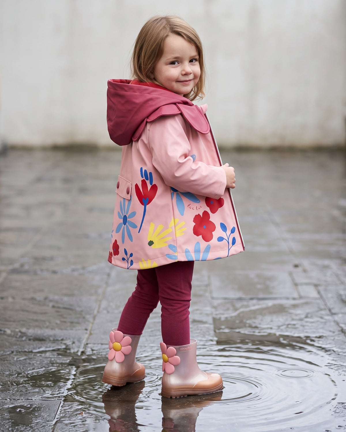 Mini Melissa kız çocuk yağmur çizmesi giyen çocuk; pembe-bakır ombre çizme, çiçek aplike, pembe yağmurluk ve bordo tayt kombini.