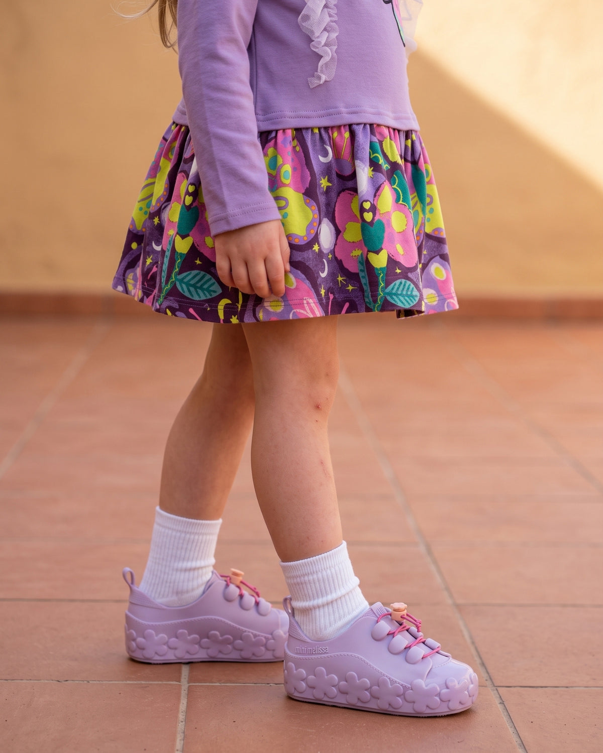 Mini Melissa lila spor ayakkabı detayı: çiçek kabartmalı taban, şeftali çiçek stoperli pembe lastik bağcık ve beyaz çorap.