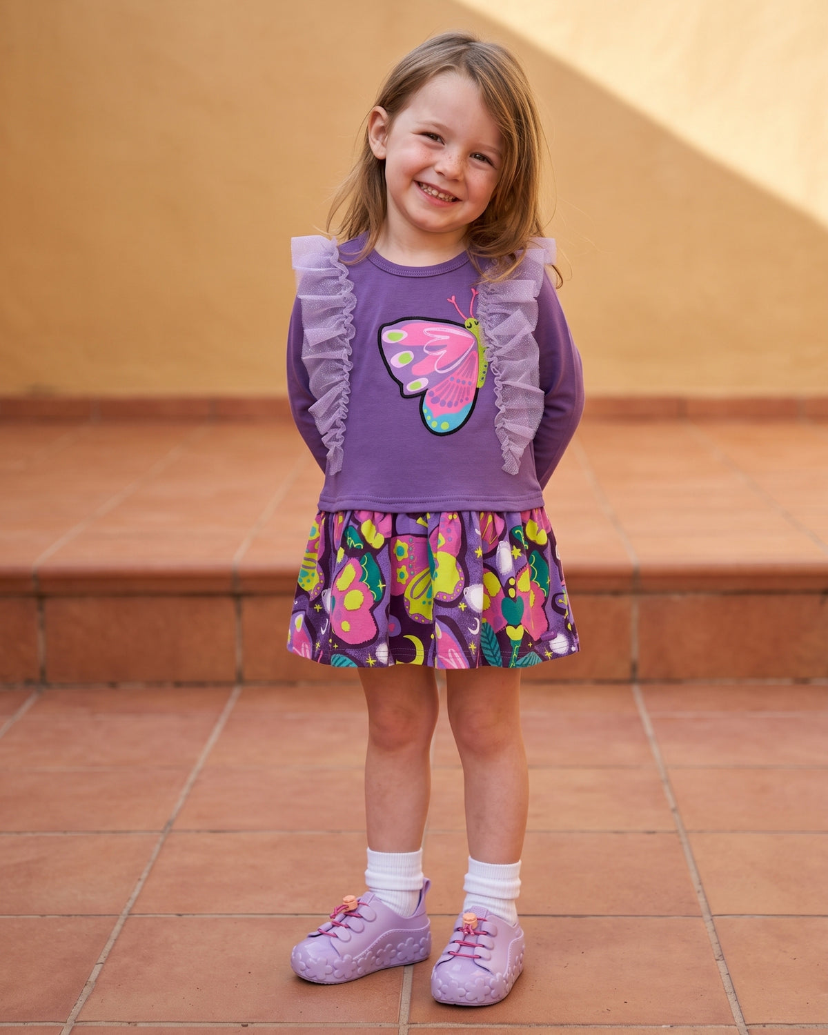 Mini Melissa mor spor ayakkabı giyen kız çocuk; lila çiçek tabanlı sneaker, kelebek aplikeli mor elbise ve beyaz çorap kombini.