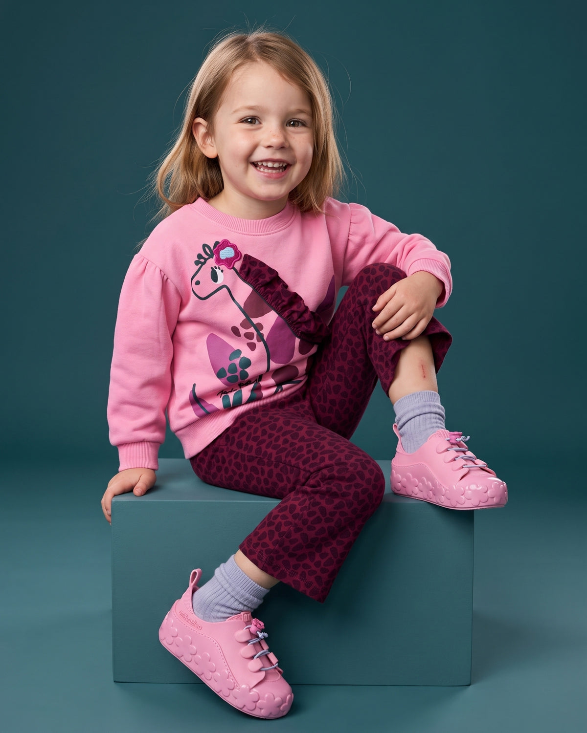 Mini Melissa pembe spor ayakkabı giyen kız çocuk; çiçek tabanlı sneaker, zürafa baskılı pembe sweatshirt ve bordo ispanyol paça takım.