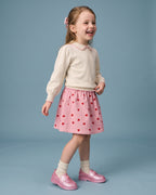 Mini Melissa pudra ayakkabı giyen kız çocuk; sedefli pembe kalp düğmeli loafer, krem triko ve kalpli pembe kadife etek kombini.