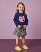 Mini Melissa sarı ayakkabı giyen kız çocuk; çiçek tabanlı sneaker, lacivert kedili sweatshirt ve gri pileli denim etek kombini.