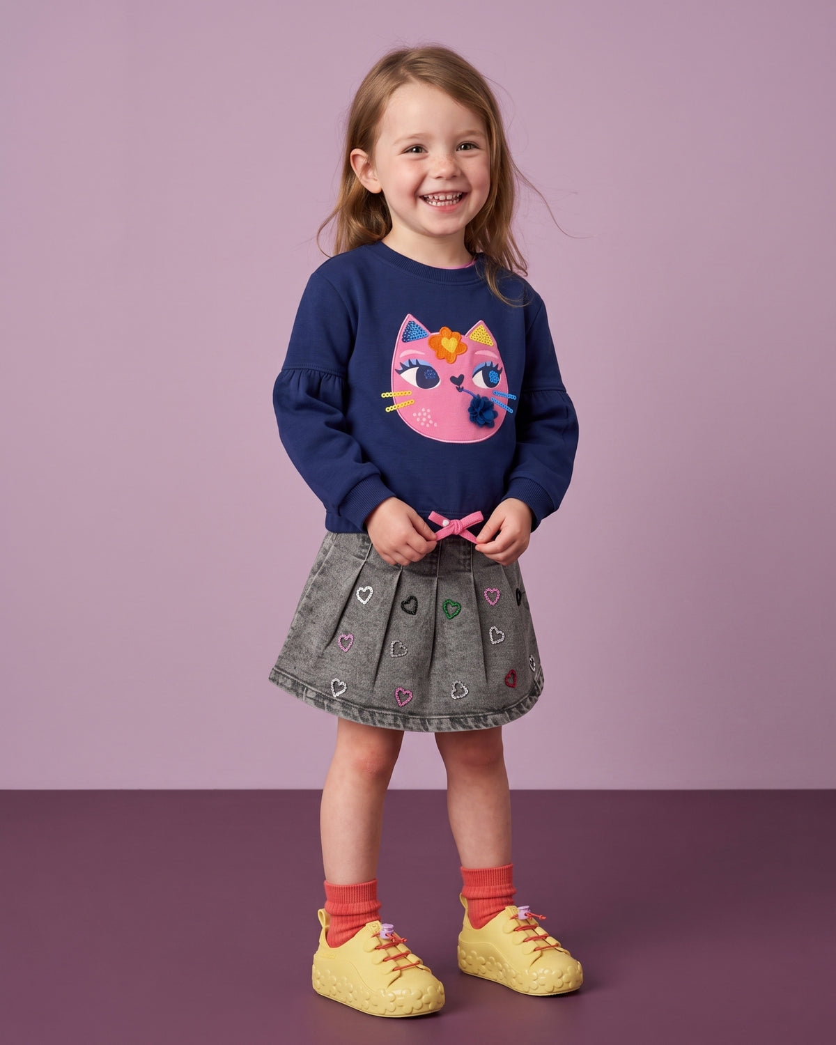 Mini Melissa sarı ayakkabı giyen kız çocuk; çiçek tabanlı sneaker, lacivert kedili sweatshirt ve gri pileli denim etek kombini.
