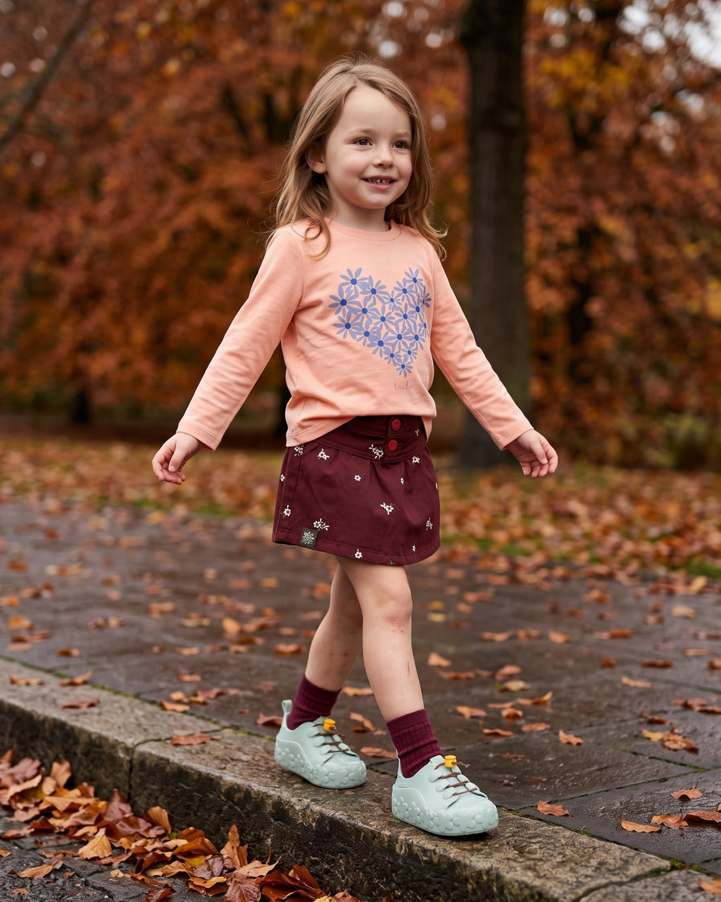 Mini Melissa yeşil ayakkabı giyen kız çocuk; su yeşili çiçek tabanlı sneaker, somon çiçekli tişört ve bordo şort etek kombini.