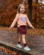 Mini Melissa yeşil ayakkabı giyen kız çocuk; su yeşili çiçek tabanlı sneaker, somon çiçekli tişört ve bordo şort etek kombini.
