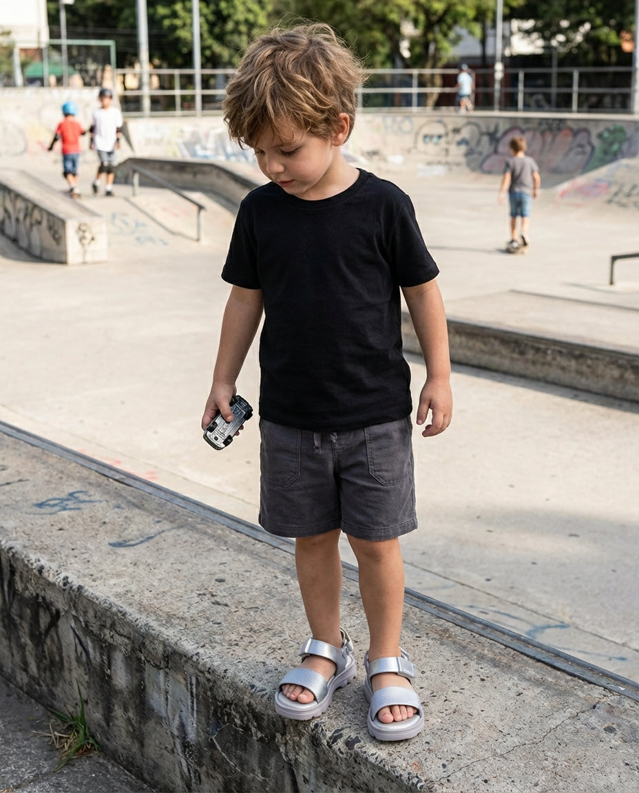 Gri şort ve siyah tişört kombiniyle sokakta duran 4 yaşındaki erkek çocuğunun ayağında kalın tabanlı gümüş Mini Melissa sandalet