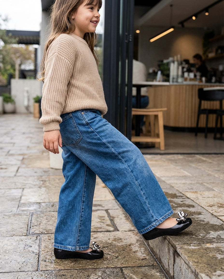 Geniş paça kot pantolonla adım atan 6 yaşındaki kızın paçasından görünen ışıltılı Mini Melissa siyah metalik fiyonklu ayakkabı