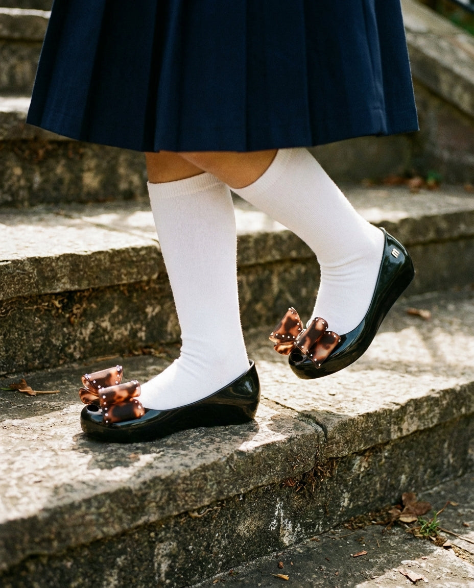 Okul forması ve beyaz dizaltı çorapla kombinlenmiş şık Mini Melissa siyah tortoise fiyonklu kız çocuk babeti