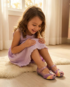 Mini Melissa Unicorn lila simli ayakkabı giyen kız çocuk, halı üstünde oturup ayakkabısına gülüyor