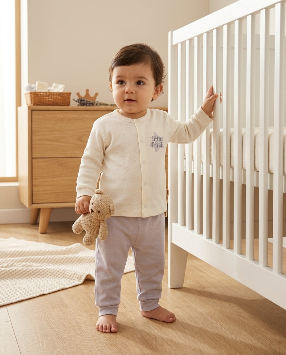 Minimalist bebek odasında beyaz karyola yanında ayakta duran Türk bebeği, uzun kollu pointel üst ve gri pantolon giymiş