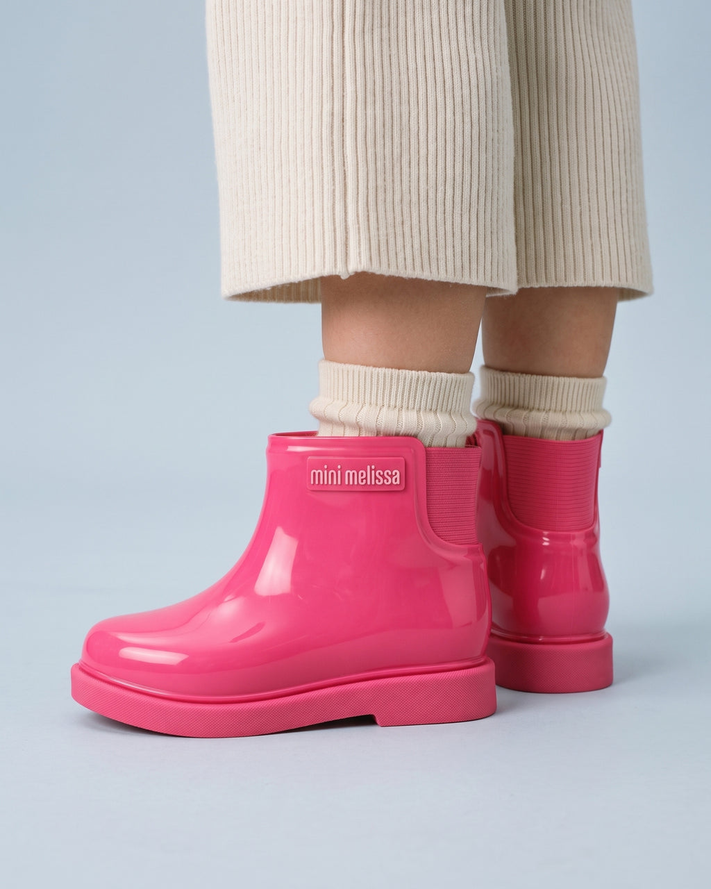 Ayakta duran kız çocuğunun ayağında Mini Melissa Chelsea pembe çocuk bot; krem ribana çorap, parlak gövdesi ve kalın mat tırtıklı tabanı yakın planda.