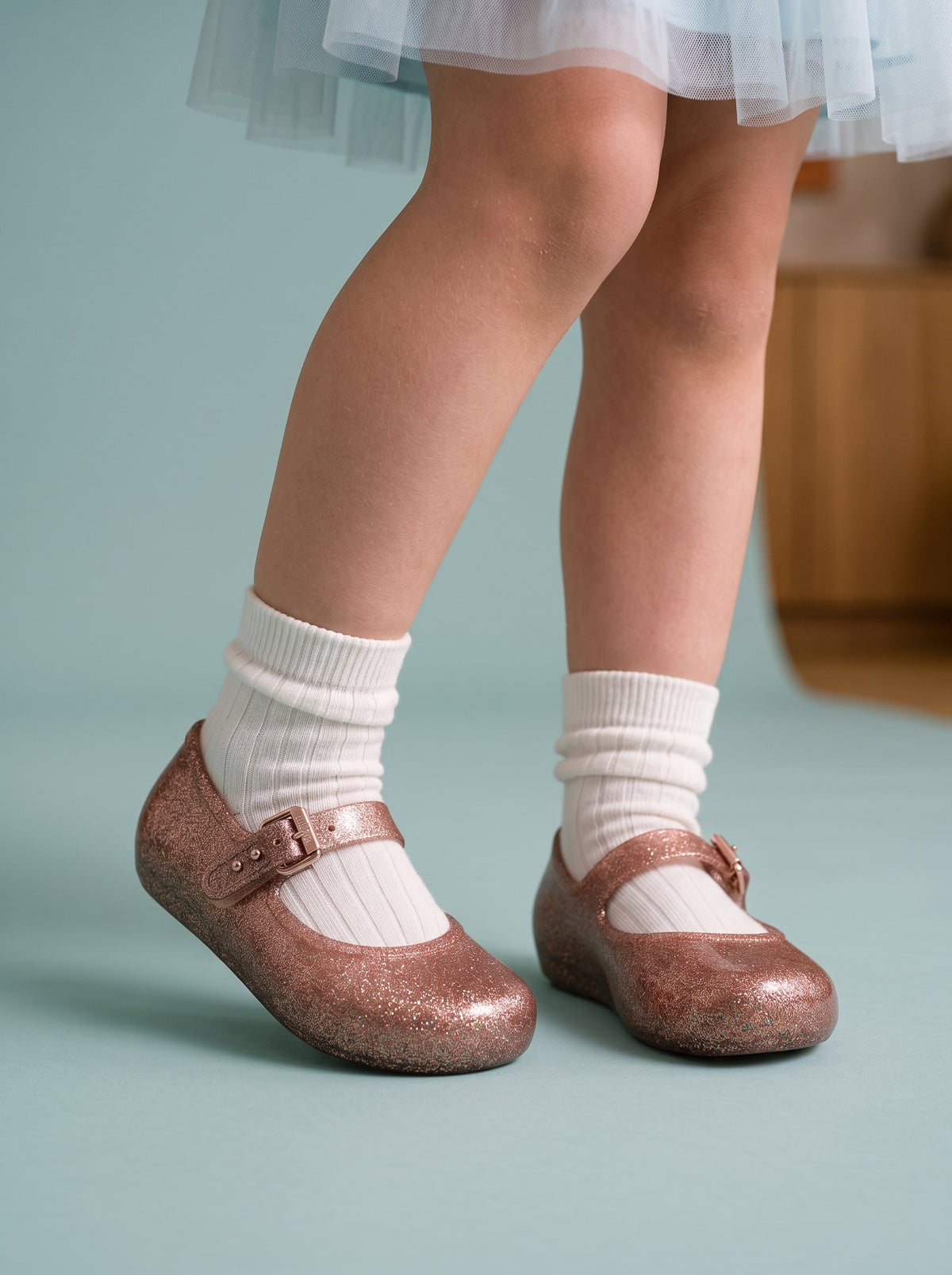 simli-Ayakta duran küçük kız çocuğunun ayağında Mini Melissa rose gold simli babet; ışıkta ayrı ayrı parlayan sim parçacıkları, tokalı bandı ve ince düz tabanı yakın planda.-kiz-cocuk-ayakta-gorunum