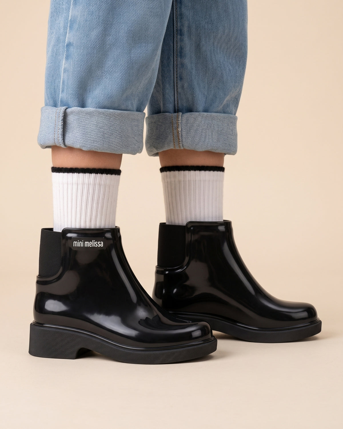 Okul çağındaki kız çocuğunun ayağında parlak siyah Mini Melissa bot; siyah çizgili beyaz çorap, kalın mat taban ve elastik yan körük yakın planda. 31-34 numara siyah okul ayakkabısı.