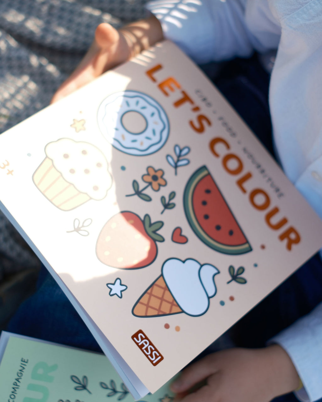 Çocuğun kucağında duran Let's Colour Food boyama kitabının kapağı